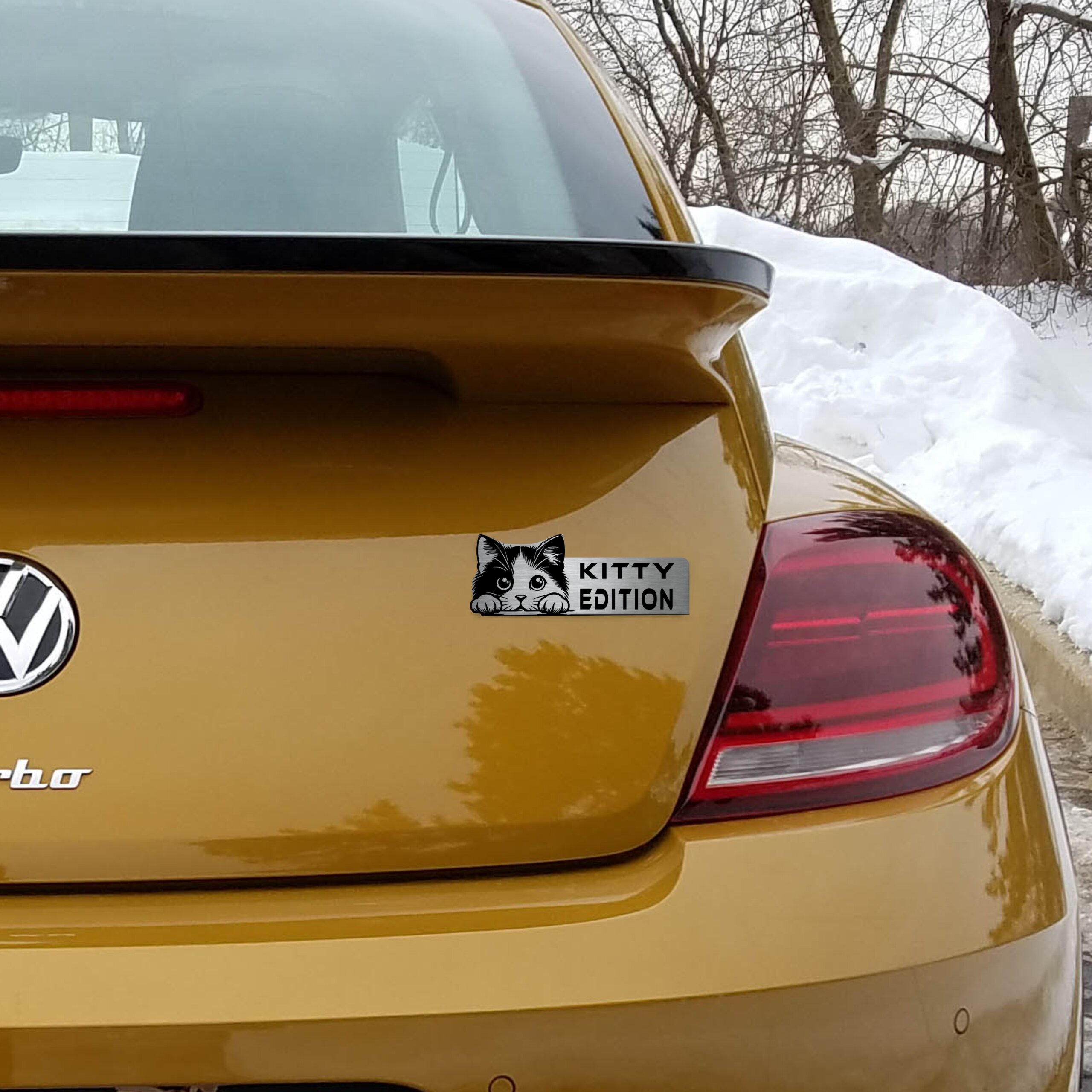 Αυτοκόλλητο Kitty Edition σε μεταλλικό φινίρισμα, με κεφαλή γάτας, τοποθετημένο στο πορτ-μπαγκάζ ενός κίτρινου αυτοκινήτου.